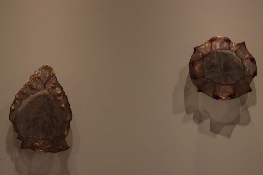 “Flora Meshell #6 (2019)” and “Flora Meshell #7 (2019)” by Lanny Bersteel hangs on the wall. It has steel mesh cut into shapes and the flame design is created with a torch, hand models and stitches.