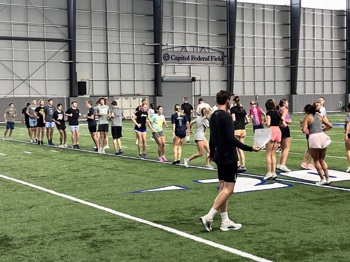 The WU cross country team engaged in warm-up exercises during an indoor practice session at the Indoor Athletic Facility. Athletes are gearing up for the upcoming season under the guidance of Myles Bach, new assistant coach.